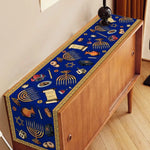Sur un meuble en bois se trouve un chemin de table bleu décoré de motifs symboliques de la culture juive, tels que des menorahs, des étoiles de David et des objets religieux.