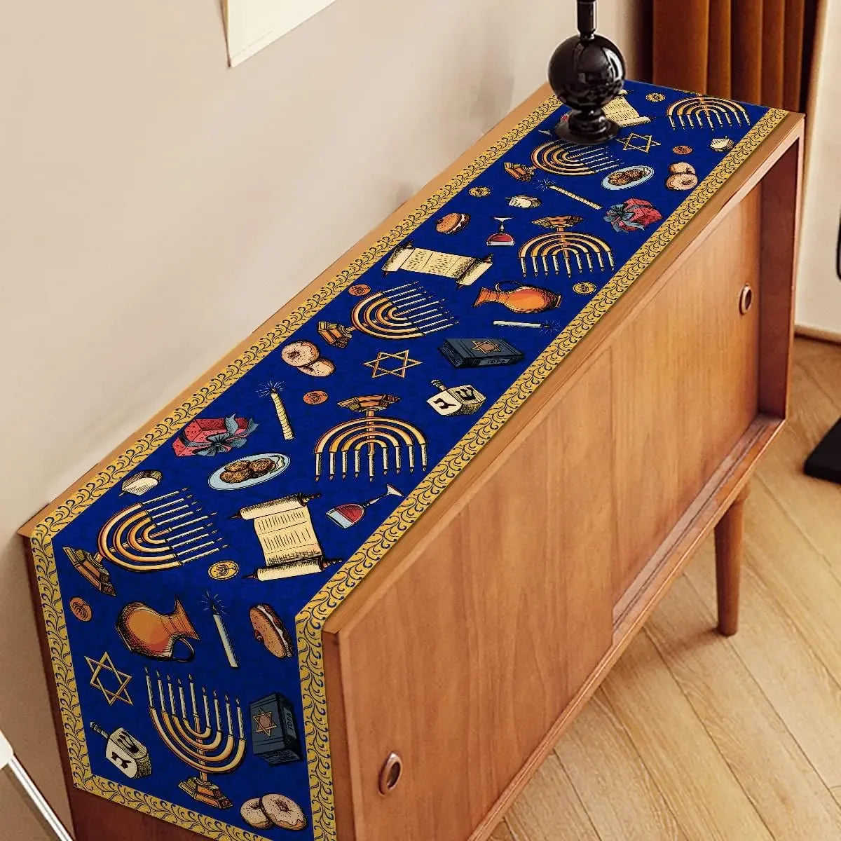 Sur un meuble en bois se trouve un chemin de table bleu décoré de motifs symboliques de la culture juive, tels que des menorahs, des étoiles de David et des objets religieux.