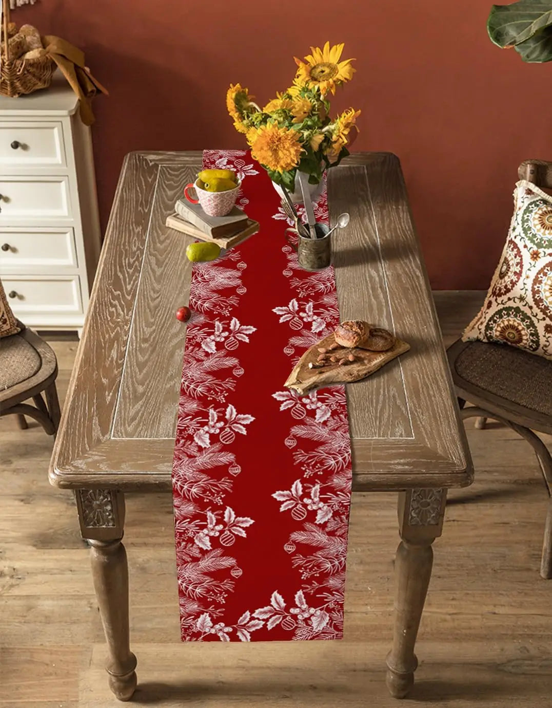 Une table en bois rustique avec un chemin de table rouge décoré de motifs floraux blancs, une bouquet de fleurs jaunes au centre et des accessoires de cuisine posés dessus.