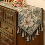 Cette image montre un chemin de table décoratif avec un motif floral élaboré, rehaussé de pompons et de glands en tissu, posé sur une commode en bois vintage. Il crée une ambiance chaleureuse et ornementée dans cette pièce.