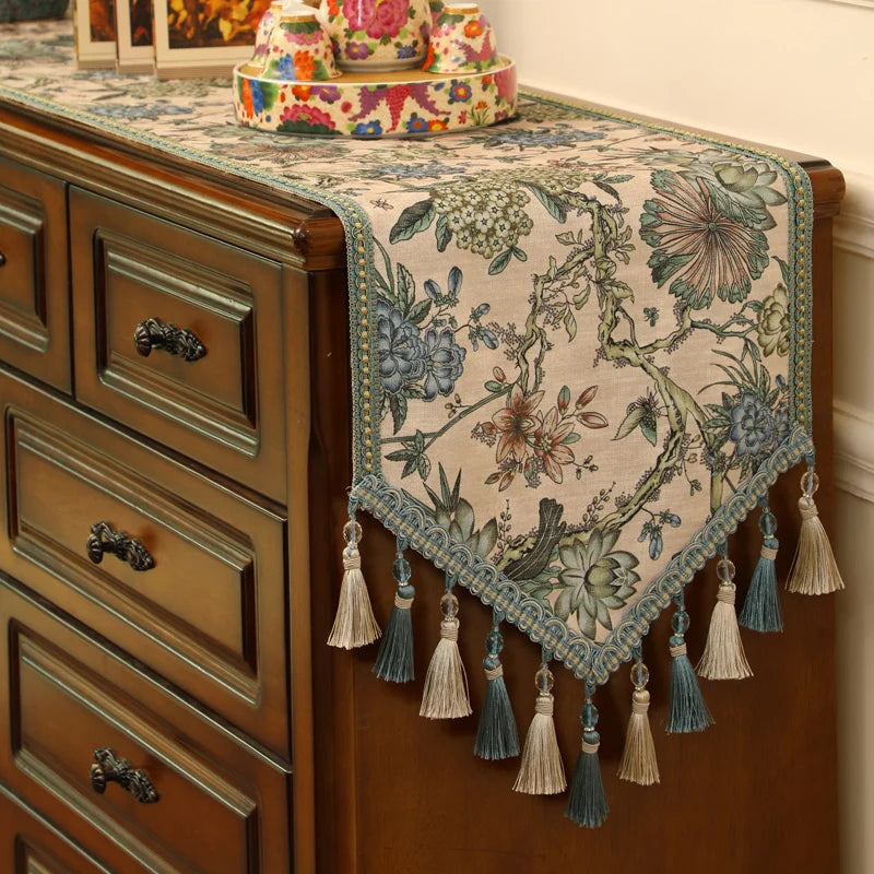 Cette image montre un chemin de table décoratif avec un motif floral élaboré, rehaussé de pompons et de glands en tissu, posé sur une commode en bois vintage. Il crée une ambiance chaleureuse et ornementée dans cette pièce.