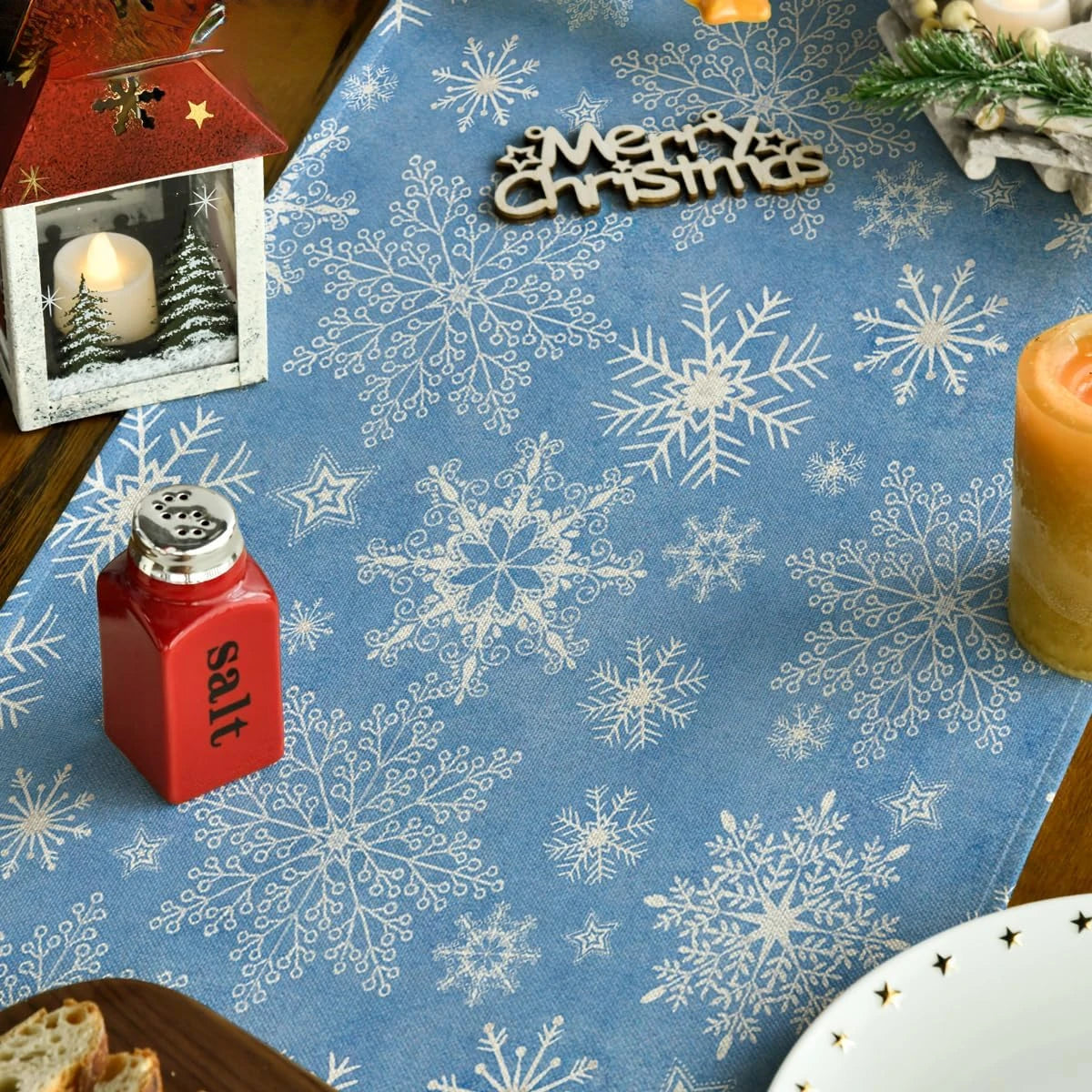 L'image montre un motif de flocons de neige blancs sur un fond bleu, ainsi qu'un petit cadre contenant une bougie allumée et du texte "Merry Christmas" en bois. Il y a également un pot de sel de cuisine et un décor de bougies et d'éléments floraux sur un plan de travail en bois.