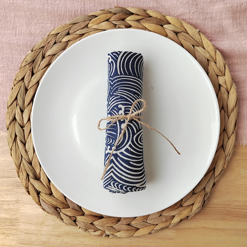 Une serviette de table bleue et blanche enroulée avec de la ficelle sur une assiette blanche posée sur un dessous de plat en osier.