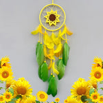 Un capteur de rêve décoratif avec un grand tournesol au centre, entouré de plumes jaunes et vertes, suspendu dans un cercle doré. Autour de l'objet, on voit de magnifiques tournesols jaunes.