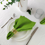 Cette image met en scène une décoration de table élégante avec une serviette en tissu vert, une assiette blanche, une fourchette et un couteau sur une nappe blanche, complétée par des éléments décoratifs tels qu'une bougie et un verre en cristal.