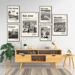 Une série d'affiches évoquant différentes villes américaines, encadrées sur un mur, avec un meuble en bois clair et des plantes d'intérieur.