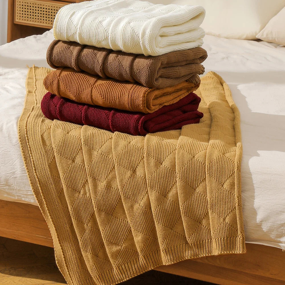 Cette image montre une pile de serviettes et de couvertures pliées de différentes couleurs, telles que l'ivoire, le brun et le bordeaux, sur un lit.