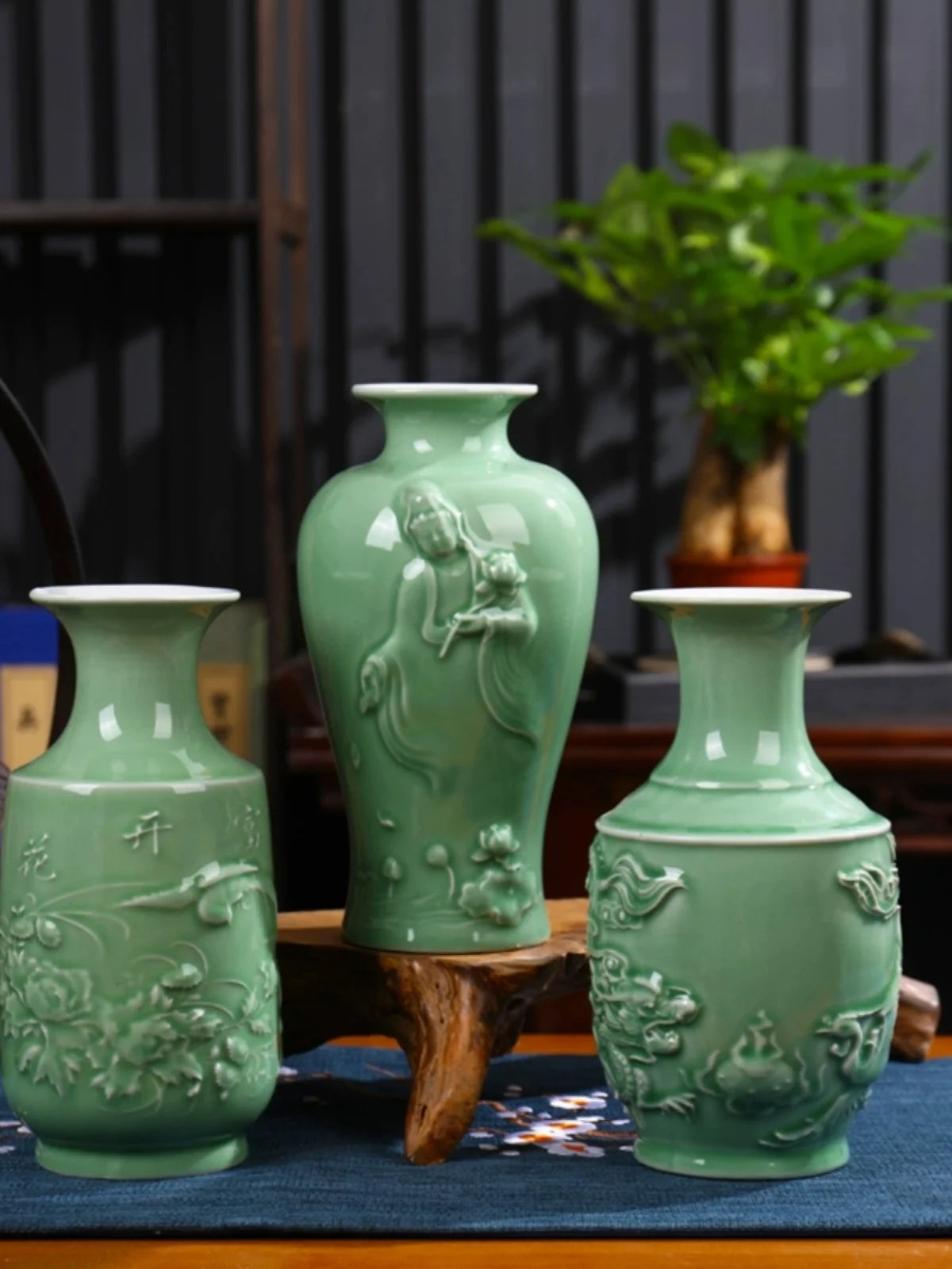 Élégants vases en céramique vert clair ornés de motifs floraux et figuratifs, disposés sur une table avec une plante en pot.