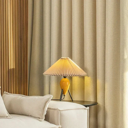 Lampe de table design moderne avec abat-jour plissé, posée sur une table dans un salon élégant avec rideaux beiges. Ambiance chaleureuse et intime pour une décoration intérieure raffinée. Luminaire idéal pour un éclairage d'appoint dans un espace de vie contemporain et stylé.