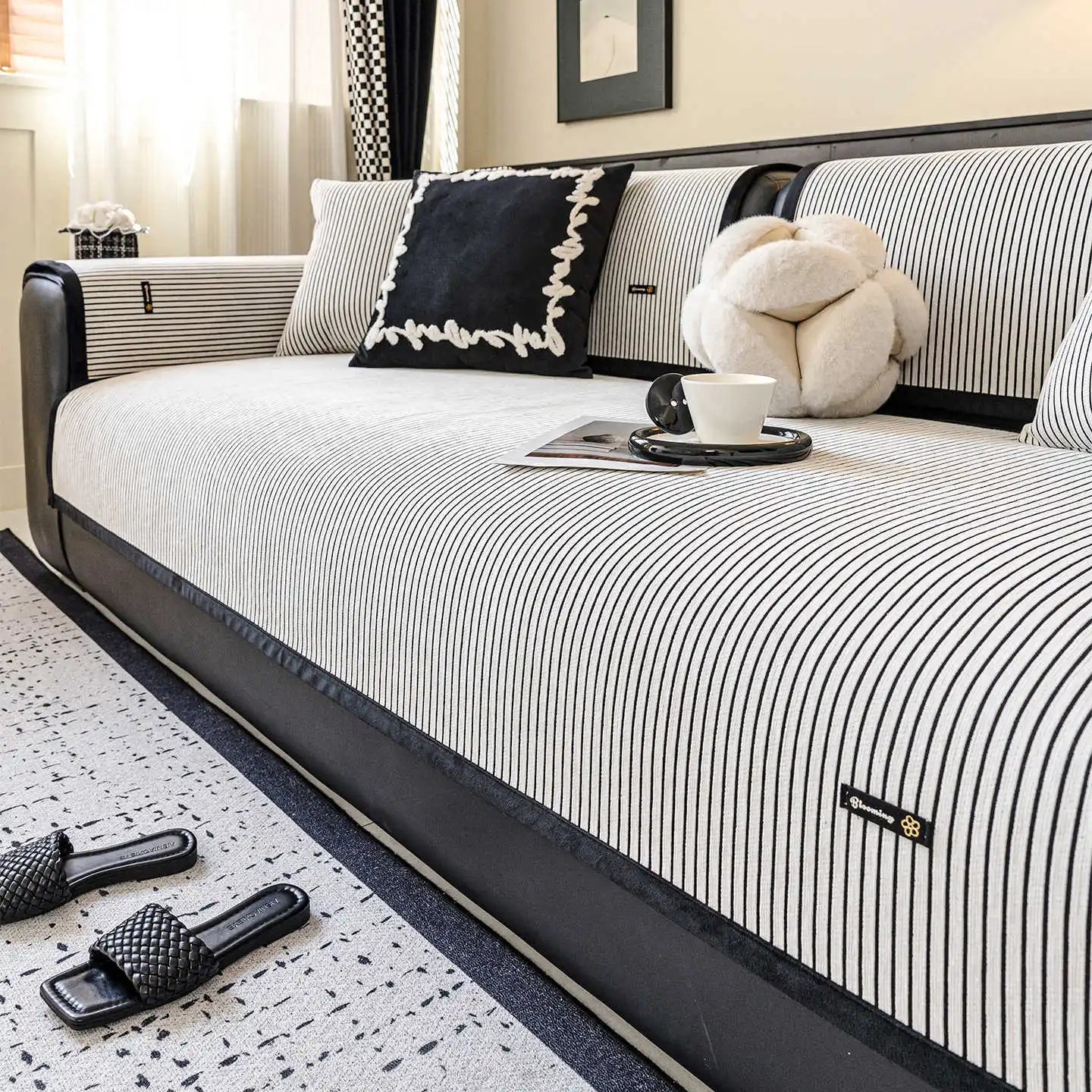 L'image montre un lit avec un cadre rayé noir et blanc, des coussins décoratifs et une tasse de café sur un plateau à côté du lit, avec des sandales noires au sol.