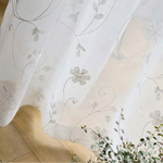 Rideaux blancs avec broderies de géraniums suspendus à une fenêtre, une nappe blanche sur la table en toile de fond et des draps de lin éparpillés autour, créant une ambiance chaleureuse et accueillante. Cette image illustre parfaitement nos gammes de rideaux et de linge de maison aux motifs floraux.