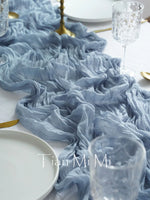 Cette image présente une décoration de table élégante, avec des tissus bleu-gris chiffonnés, une assiette blanche et un verre de cristal, suggérant une atmosphère raffinée et sophistiquée.