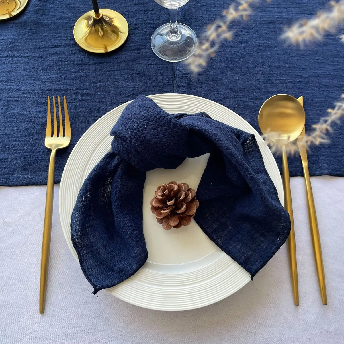 Un couvert de table élégant avec une assiette blanche, des couverts dorés, une serviette de table bleue marine et un décor de pomme de pin sur un fond bleu.