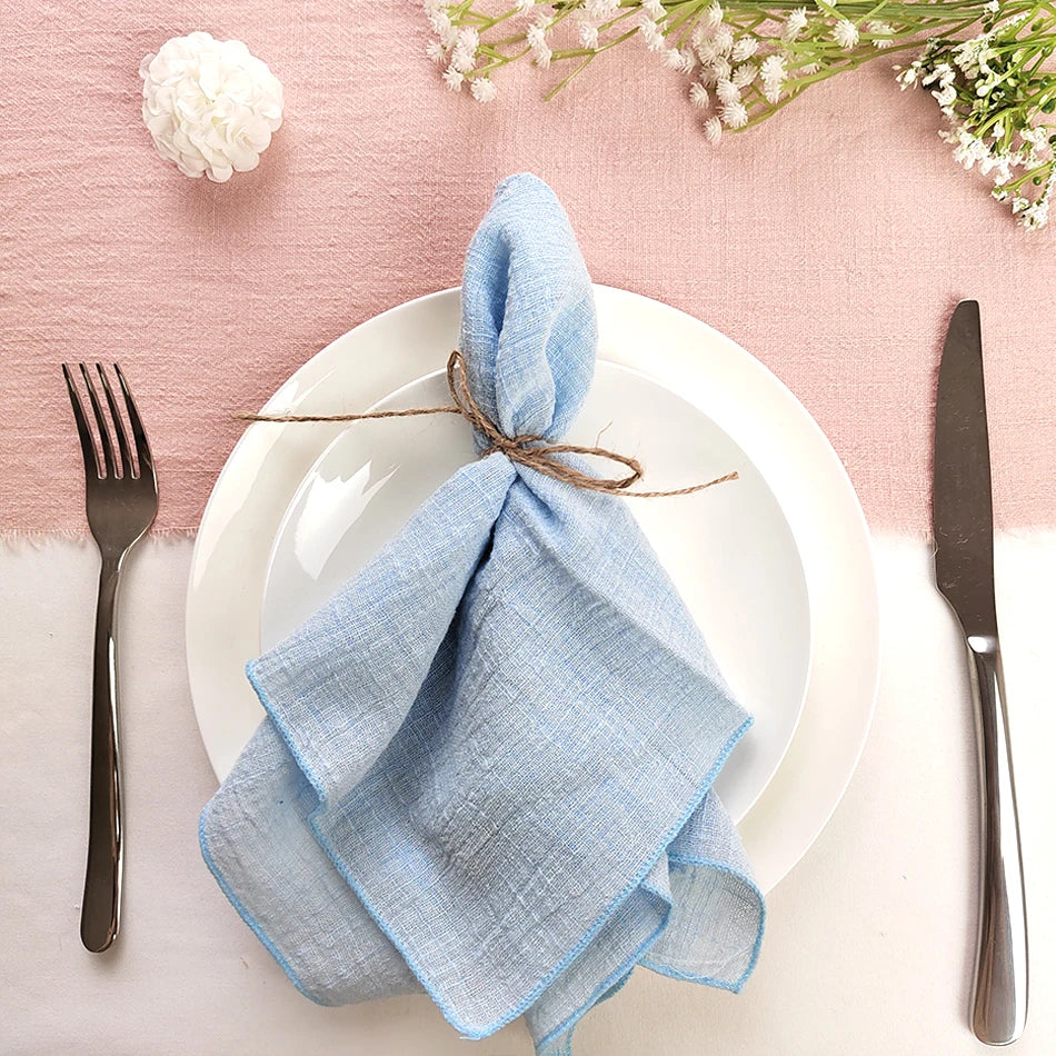 Une table élégante avec une assiette blanche, des couverts en métal et une serviette bleue pliée, le tout sur un fond rose pastel.