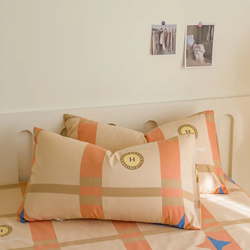 Deux coussins carrés beiges et orange, ornés d'un motif géométrique et de l'emblème de la marque, sont posés sur un lit dans une chambre minimaliste.