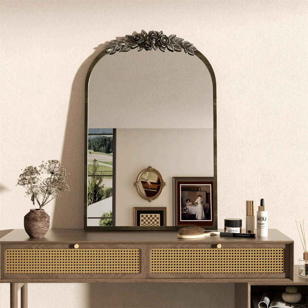 Un miroir orné de fleurs de style baroque surplombe une console en bois avec des tiroirs en cannage, sur laquelle sont posés divers objets décoratifs, encadrant une fenêtre offrant une vue sur un paysage verdoyant.