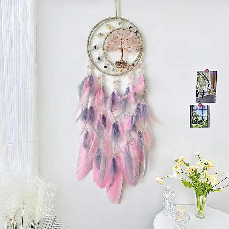 Suspension décorative en forme de cercle avec un arbre de vie en pierres roses et un attrape-rêves aux plumes roses et violettes, suspendu devant un mur blanc avec des photographies encadrées.