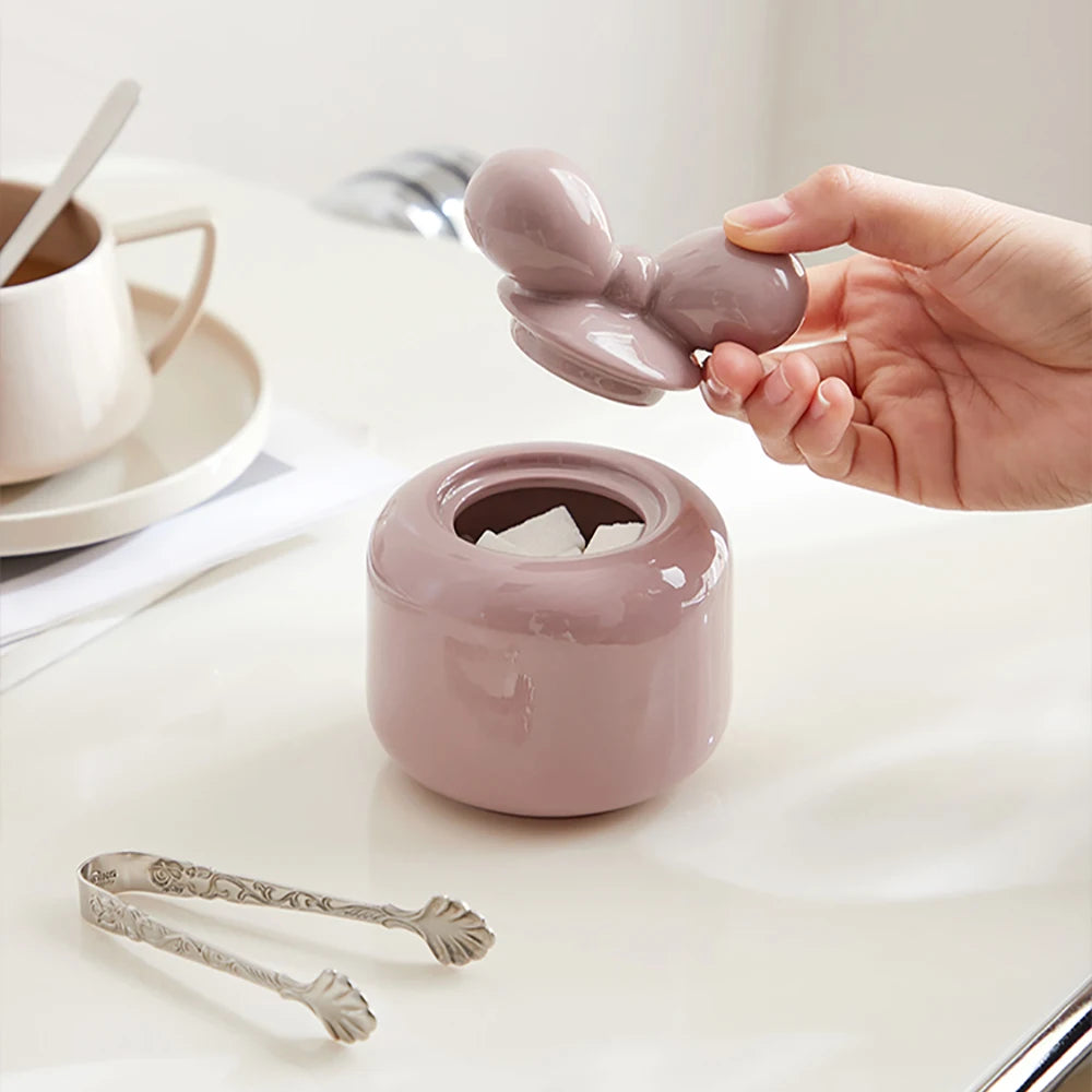 Sur la table blanche, on voit un bocal en céramique rose entouré de cuillères vintage, accompagné d'une tasse de café et d'autres accessoires de cuisine.