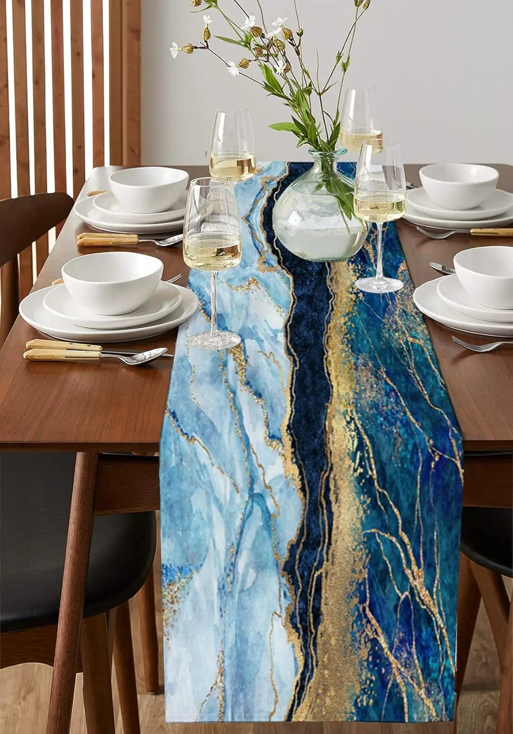 Une table élégante dressée avec des plats blancs, des verres de vin, un bouquet de fleurs sauvages et un chemin de table bleu et or évoquant le marbre.