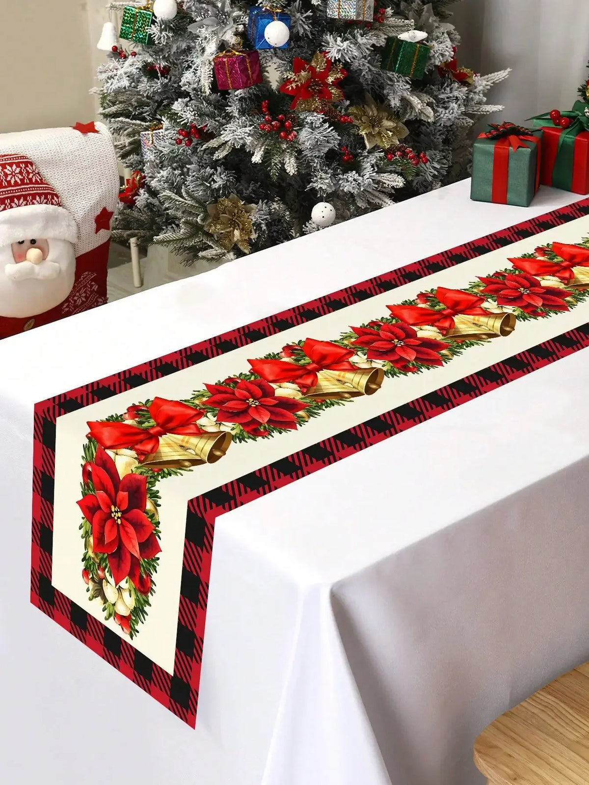 Une belle nappe de Noël décorée de motifs de poinsettias rouges, de cloches dorées et de rayures noires et rouges, dans un beau décor de Noël avec un arbre enneigé et des cadeaux.