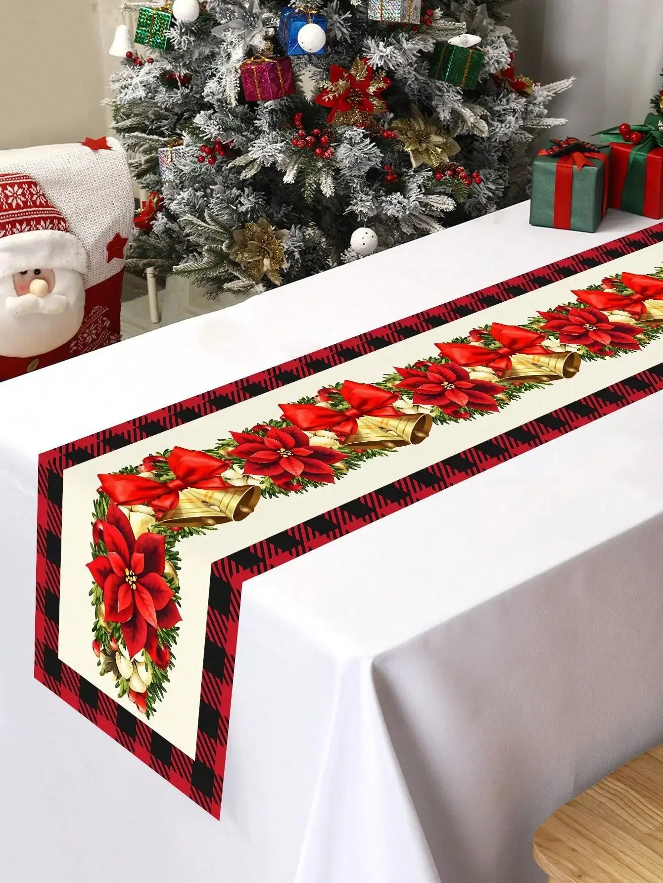 Une belle nappe de Noël décorée de motifs de poinsettias rouges, de cloches dorées et de rayures noires et rouges, dans un beau décor de Noël avec un arbre enneigé et des cadeaux.