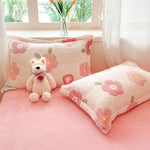 Coussins décoratifs rose pâle avec motifs de fleurs brodées, placés sur un canapé près d'une fenêtre avec des rideaux blancs, accompagnés d'un ours en peluche rose.