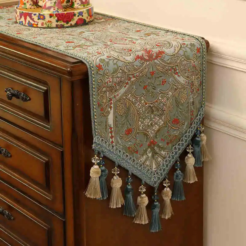 Un élégant chemin de table aux motifs floraux colorés, agrémenté de glands et de pompons décoratifs, qui confère une ambiance chaleureuse et raffinée à cette commode en bois.