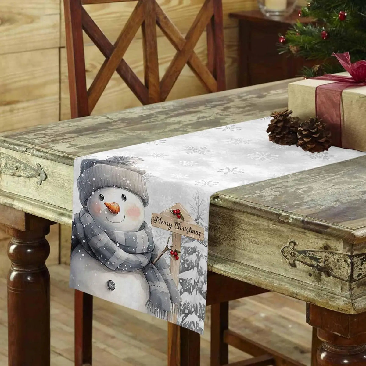 Une table en bois vintage est décorée pour Noël, avec un chemin de table orné d'un adorable bonhomme de neige en gris et blanc tenant une pancarte "Joyeux Noël".
