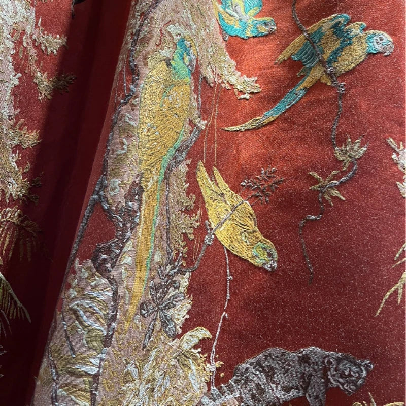 Détail du motif brodé du rideau : perroquets jaunes et verts perchés sur des branches feuillues, avec texture en relief sur fond rouge brique.
