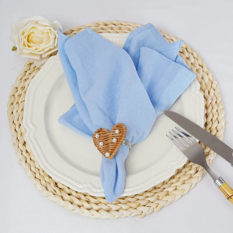 Une serviette bleue en forme de noeud avec un coeur en osier décoratif sur une assiette blanche au centre d'un set de table en fibres tressées.