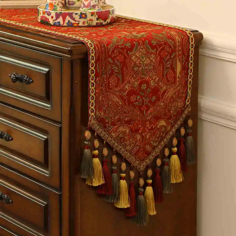 Élégant chemin de table en velours rouge orné de motifs dorés et de glands frangés, posé sur un meuble en bois.
