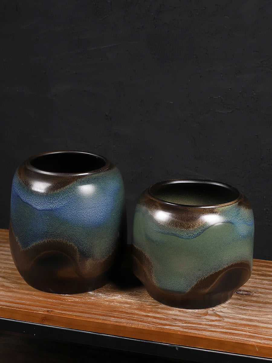 Deux vases en céramique avec un motif abstrait de couleurs bleues, vertes et brunes sur un fond noir mat, posés sur un présentoir en bois.