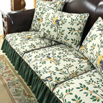 L'image montre un canapé en cuir noir avec des coussins ornés d'un motif de fleurs et de feuilles vertes, jaunes et noirs, créant une ambiance chaleureuse et naturelle dans la pièce.