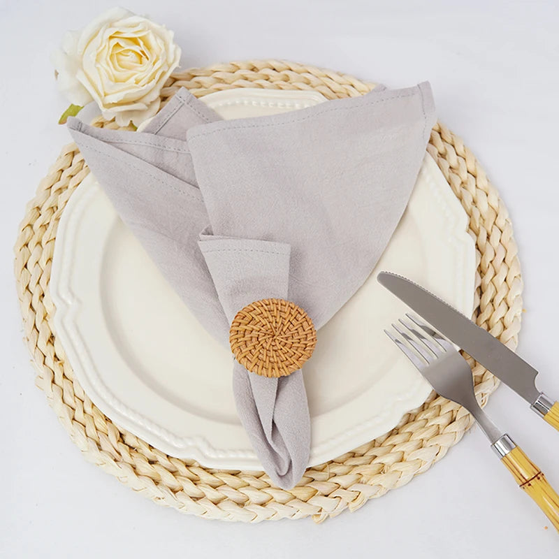 L'image montre une table de service élégante avec une assiette blanche, des couverts en métal, un napperon tressé et une serviette de table beige ornée d'un bouton en osier, ainsi qu'une belle rose jaune sur le côté.