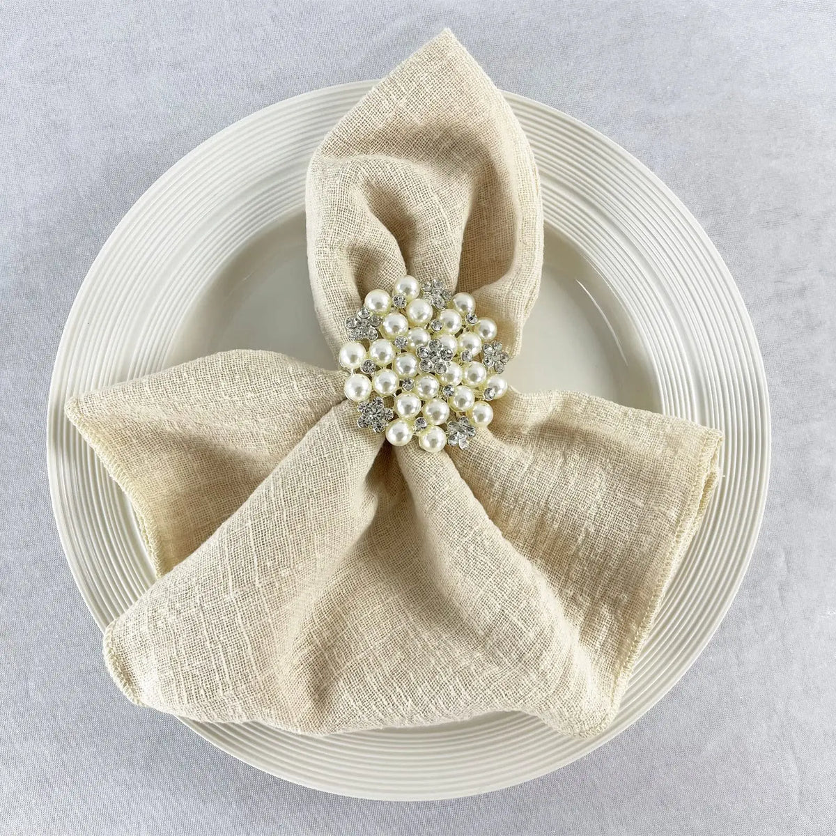 Une serviette beige en tissu avec un magnifique broche de fleurs blanches et de cristaux, élégamment disposée sur une assiette blanche.