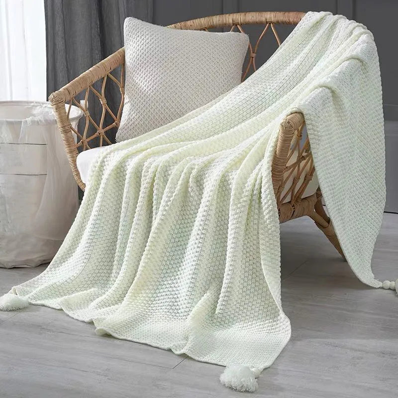 Cette image présente un fauteuil en osier avec un plaid blanc douillet et une taie d'oreiller assortie, créant une ambiance chaleureuse et cocooning dans un intérieur aux teintes neutres.