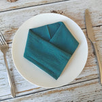 Arrangement artistique de serviette en papier bleu canard pliée sur une assiette blanche, placée sur une table en bois rustique avec des couverts.