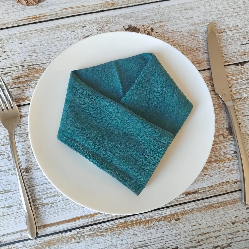 Arrangement artistique de serviette en papier bleu canard pliée sur une assiette blanche, placée sur une table en bois rustique avec des couverts.