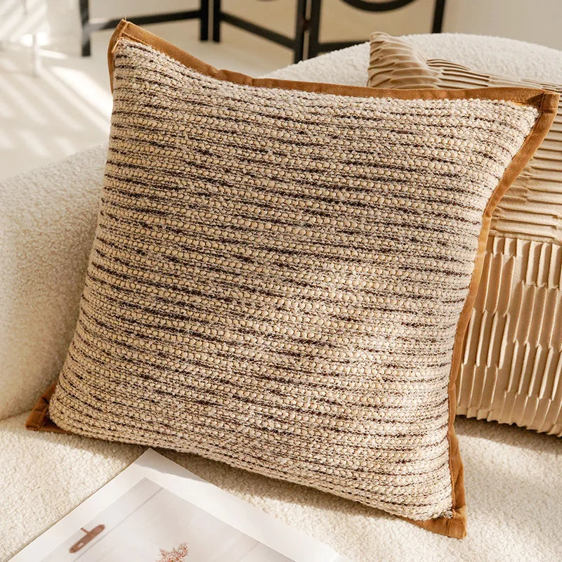 L'image montre un coussin décoratif en crochet avec des rayures horizontales beige et brun foncé, reposant sur un canapé blanc avec un autre coussin décoratif aux motifs froissés.