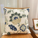Coussin décoratif brodé avec des motifs floraux vintage bleu marine, crème et beige sur un fond blanc cassé.