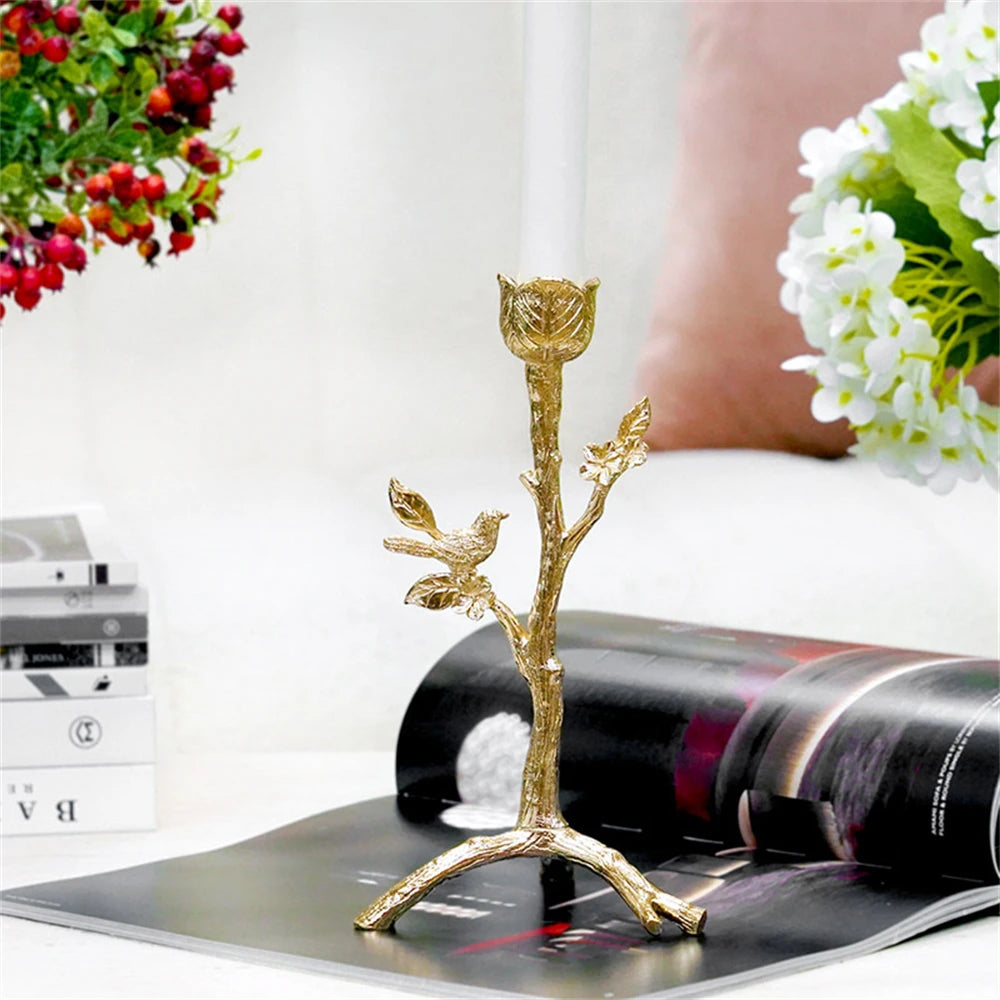 Un support décoratif en métal doré en forme de branche ornée de feuilles et d'un petit oiseau, posé sur une surface réfléchissante à côté d'une bouteille de vin et de fleurs blanches.