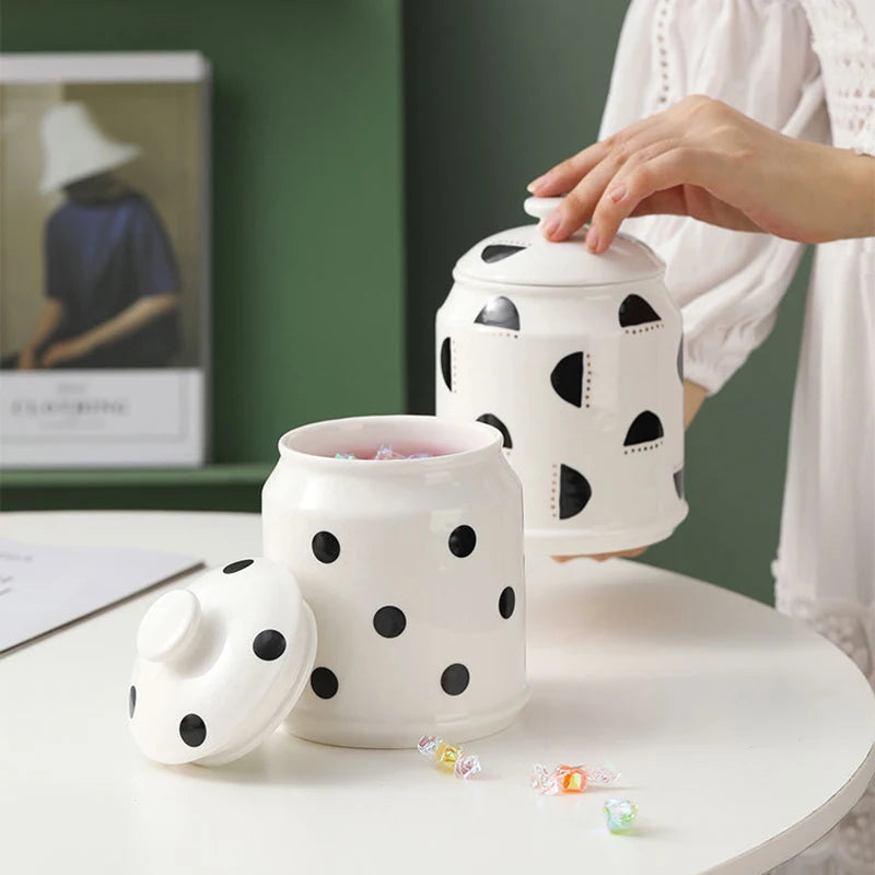 Des bocaux en céramique blanc à pois noirs, avec couvercles assortis, posés sur une table blanche, dans un intérieur moderne avec un mur vert.