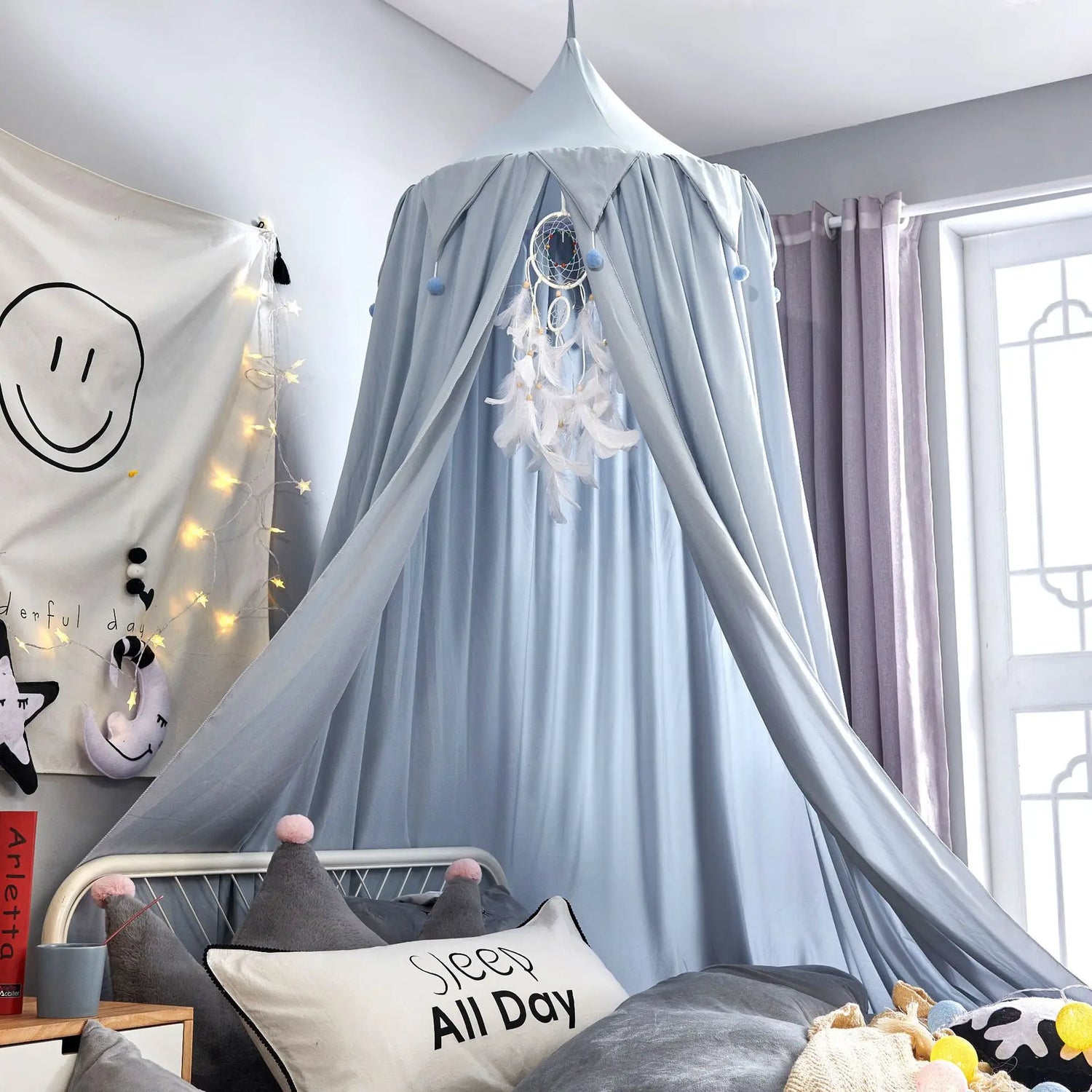 La chambre présente un lit avec un baldaquin gris clair, décoré de guirlandes lumineuses et d'un attrapeur de rêves, créant une ambiance douce et confortable.