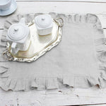 Sur une table en bois blanche, un service à thé vintage blanc et argenté avec une théière, une tasse et un plat décoratif, le tout sur un napperon en lin gris.