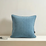 Coussin bleu pétrole avec motif à rayures verticales, posé sur une surface blanche avec un vase en verre.