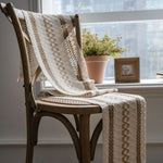 Chaise en bois avec couverture beige et motifs ethniques, placée près d'une fenêtre avec une plante verte dans un pot en céramique.