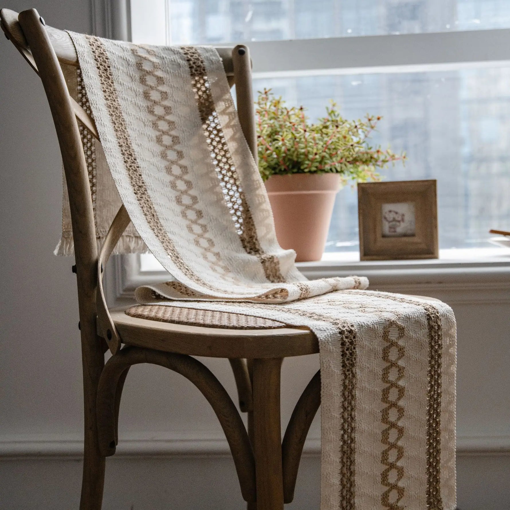 Chaise en bois avec couverture beige et motifs ethniques, placée près d'une fenêtre avec une plante verte dans un pot en céramique.