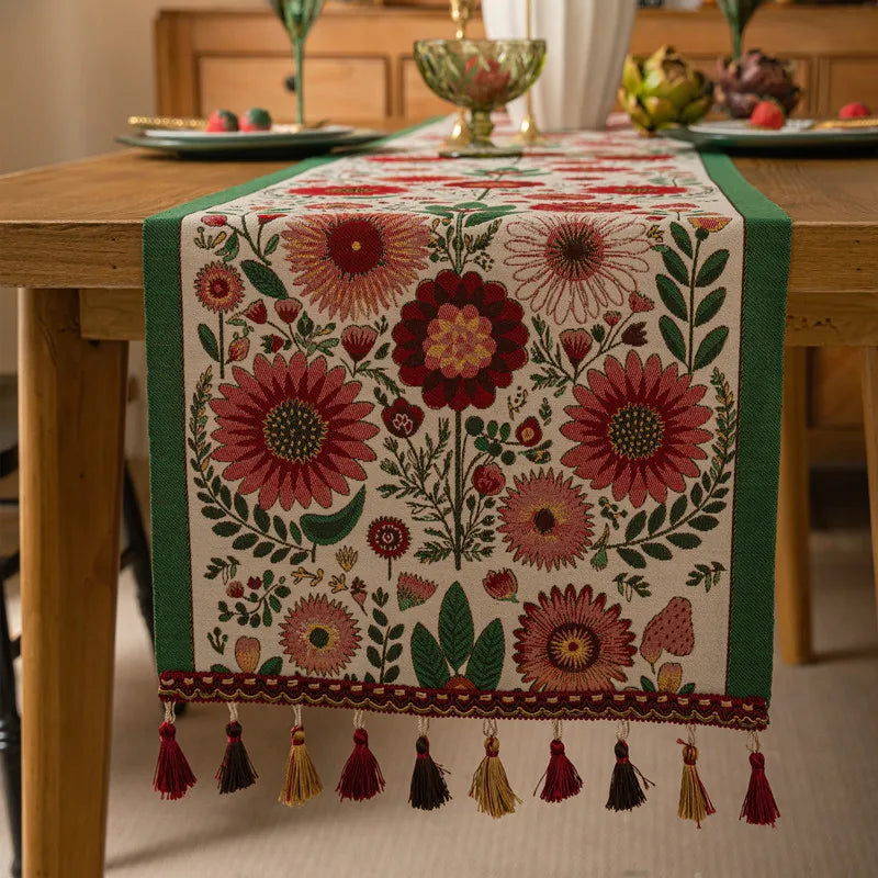 Un chemin de table floral richement décoré avec des motifs de fleurs rouges et vertes sur un fond clair, avec des glands aux extrémités, créant un look élégant et chaleureux pour une table de salle à manger.