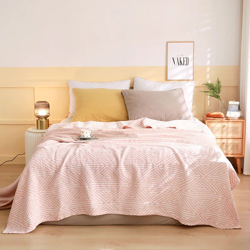 Cette chambre à coucher confortable et bien aménagée présente un lit avec une couverture rose pastel aux motifs géométriques, des coussins gris et beiges, une lampe de chevet dorée et un cadre mural avec l'inscription "Naked", créant une atmosphère chaleureuse et apaisante.