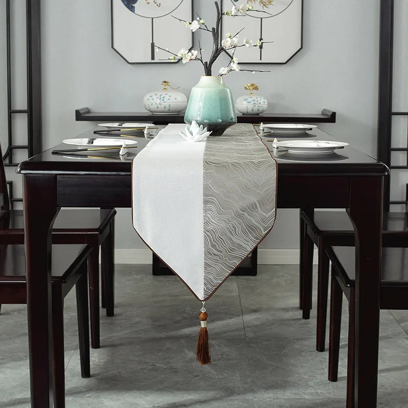 Cette image montre une table de salle à manger élégante, avec un chemin de table gris orné d'un vase bleu aqua et de fleurs de cerisier blanches, encadrée par des chaises noires et des éléments décoratifs muraux en noir et blanc.